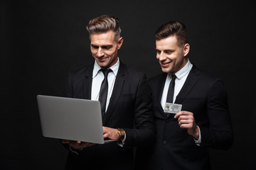 Poster - Handsome excited happy two business men posing isolated over black wall background using laptop computer holding credit card.