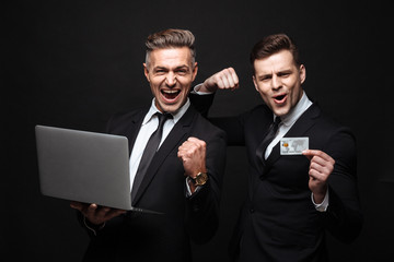 Handsome excited happy two business men posing isolated over black wall background using laptop computer holding credit card.