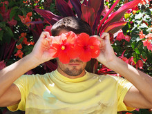 Portrait Of An Adult Man Wearing A Yellow T-shirt In A Tropical Garden Holding Red Hibiscus Flowers In Place Of His Eyes.