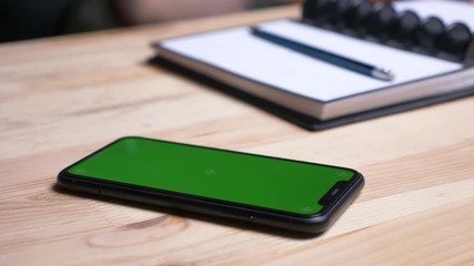 Closeup shoot of female hand taking a phone with green screen laying on the desk next to the notebook indoors in the office