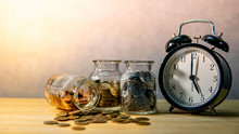 Clock With Coins In Currency Glass Jars And Spilling On Wooden Table. Saving Money For Future Retirement. Financial Business Growth. Time Investment Concept