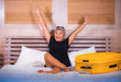 © TheVisualsYouNeed - Attractive and happy mature Asian woman smiling cheerful arriving at hotel room sitting on bed with suitcase enjoying holidays or business trip