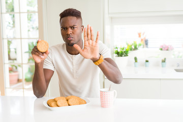 Sticker - African american man eating healthy whole grain biscuit with open hand doing stop sign with serious and confident expression, defense gesture