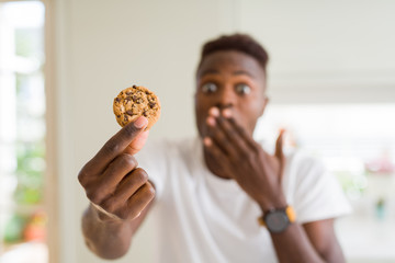 Poster - Young african american man eating chocolate chips cookies cover mouth with hand shocked with shame for mistake, expression of fear, scared in silence, secret concept