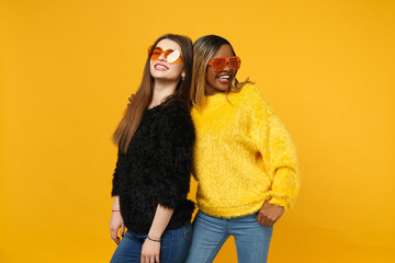 Two young women friends european and african american in black yellow clothes standing posing isolated on bright orange wall background, studio portrait. People lifestyle concept. Mock up copy space.