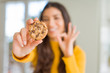© Krakenimages.com - Young woman eating chocolate chips cookies at home doing ok sign with fingers, excellent symbol