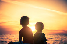 Two Boys Sitting On The Rock At The Beach At Sunset