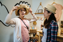 Girlfriends On Shopping Spree Trying Ladies Hats And Fashion Items In Accessories Store. Two Asian Women Shopaholics Wear Straw Hat Looking At Mirror In Small Village. Travel Purchase Lifestyle.