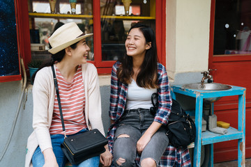 Wall Mural - group of asian girlfriends sitting on bench talking relax in spring sunny day. happy women tourists laughing outside little shop selling ice cream by sink to wash hands in small village travel.