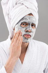 man in bathrome touching facial mask and looking at camera isolated on grey