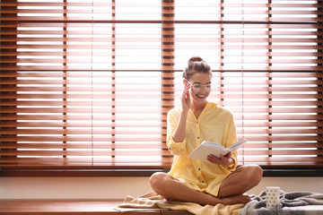 Canvas Print - Young woman reading book near window with blinds at home. Space for text