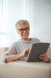 © DragonImages - Portrait of mature woman with short hair wearing eyeglasses working online on digital tablet while sitting on sofa at home