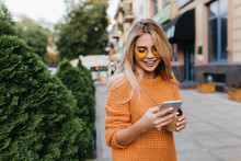 Lovely Blonde Girl Walking By Green Bushes With Smile, Carrying Smartphone And Cup Of Coffee. Stunning Fair-haired Woman In Yellow Attire Hurrying To Somewhere And Drinking Latte.
