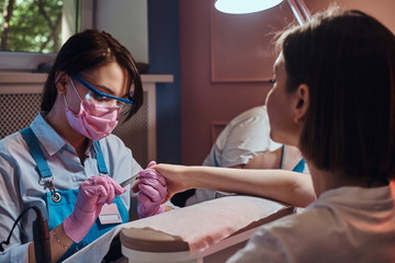 Wall Mural - Talented dilligent manicurist in protective mask is working on customer nails with nail file.