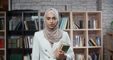 Wall Mural - Portrait of female muslim student, wearing hijab, who is at campus library and confidently looking at camera - modern islam concept 4k