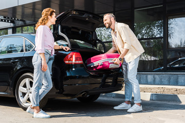 Wall Mural - low angle view of happy man putting pink luggage in car trunk near woman