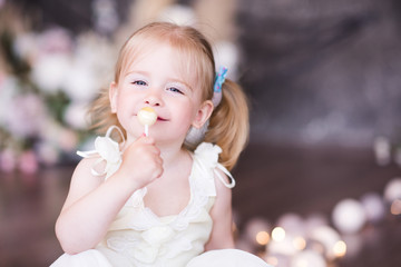 Wall Mural - Smiling baby girl 1-2 year old holding lollipop posing in room close up. Looking at camera. Childhood.