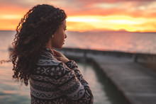 Thoughtful Evening Mood With A Young Afro American Woman Standing On The Promenade By The Lake And Looking Towards The Water And The Setting Sun. Burning Sky