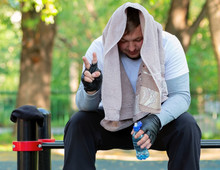 A Young Guy In Bright Sports Clothes With A Towel On His Head And A Bottle Of Water In His Hands Is Sitting On The Gym In The Open Air. Man Resting After A Hard Workout Showing Hand Symbol Of Victory.