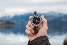 Beautiful Male Hand Holds A Magnetic Compass Against The Background Of The Mountain And A Lake. The Concept Of Finding Yourself The Way And The Truth.