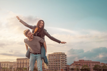 Young guy piggybacking cheerful girlfriend like airplane on urban background with sky and some buildings. Copy space in the sky.