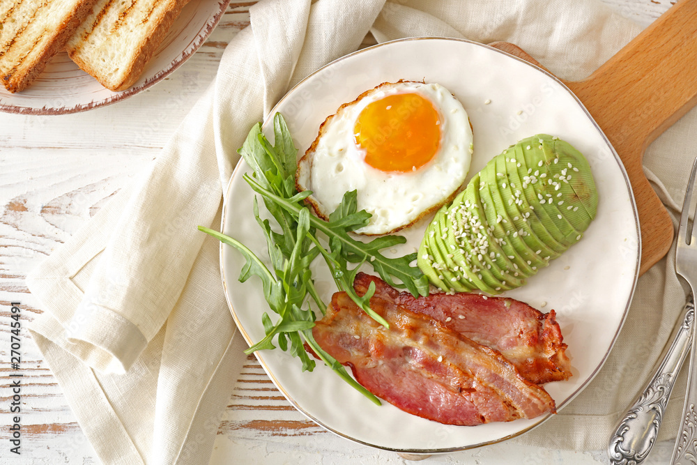 Plate with tasty fried egg, avocado and bacon on wooden table