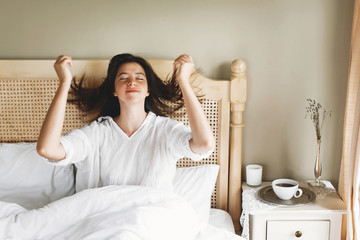 Beautiful happy young woman lying in bed in the morning in hotel room or home bedroom. Stylish brunette girl in white relaxing on white bed, waking up in soft light. Space for text