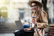 © dianagrytsku - Portrait of a glamorous young woman holding on her knees portable laptop computer while sitting on a wooden bench. Stylish female drinking coffee while relaxing after work on netbook during free time