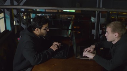 Wall Mural - tracking right of two young multiethnic men in hoodie sitting at laptops in front of each other and 