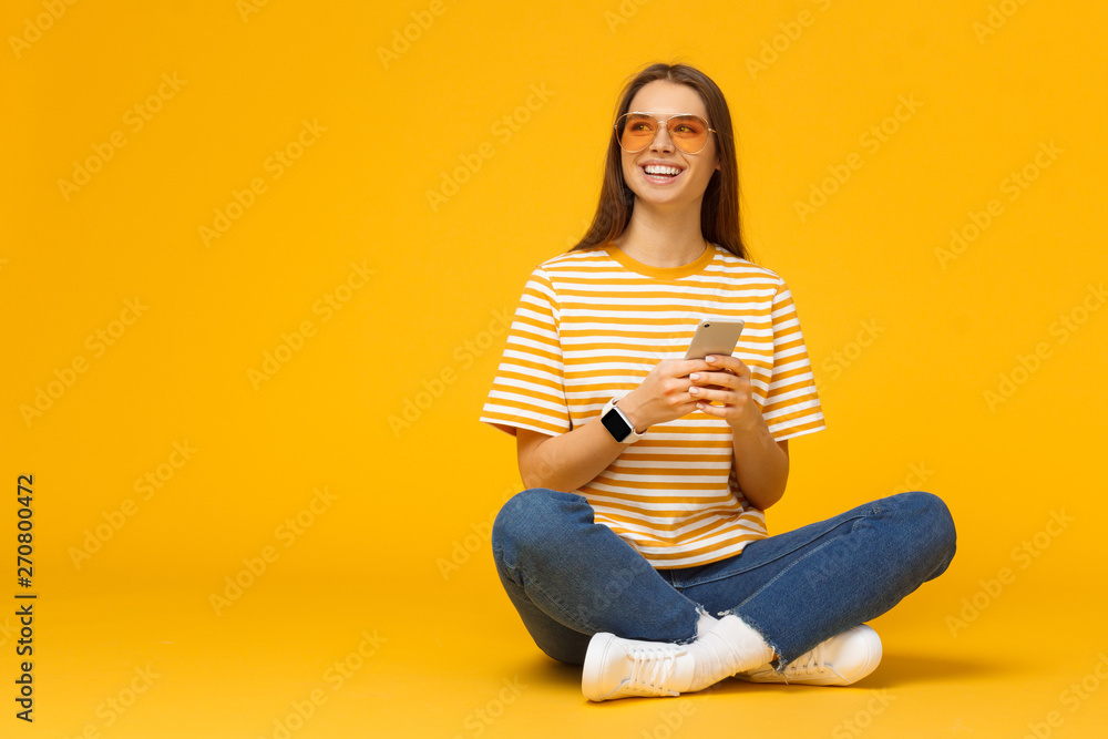 Happy young girl sitting on the floor, holding smartphone in hands and looking away, isolated on yellow background with copy space - obrazy, fototapety, plakaty 