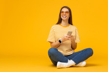 Happy Young Girl Sitting On The Floor, Holding Smartphone In Hands And Looking Away, Isolated On Yellow Background With Copy Space