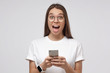 © Damir Khabirov - Wow! Close-up shot of young girl in white t-shirt, holding smartphone, looking surprised and shocked, isolated on gray background.