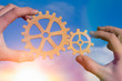 © gerasimov174 - Hands businessman holding gears on the background of the sky in the sun, connecting the puzzle pieces. creation. mechanism. Business idea concept, teamwork, strategy, innovation, cooperation