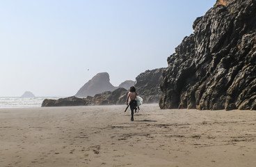 Young man surfer with no shirt and bushy long dark hair walks down misty beach with towering wet rock cliffs toward ocean with waves rolling in - shades of brown and selective focus