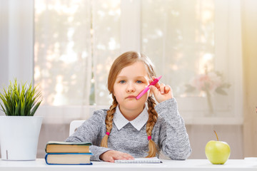 Little student girl sitting at the table. Schoolgirl with a pen in hand and a notebook, the child is thoughtful. The concept of education and school. In the background is the light from the window.