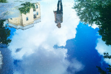 The Girl's Reflection In A Puddle On The Pavement On A Background Of White Clouds. Background With Reflections Of Clouds And A Girl In The Water.