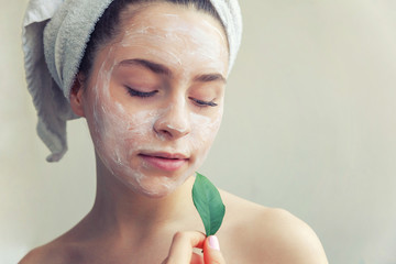 Wall Mural - Beauty portrait of woman in towel on head with white nourishing mask or creme on face and green leaf in hand, white background isolated. Skincare cleansing eco organic cosmetic spa concept