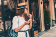canvas print picture - Young stylish woman using phone walking on the street, travel with backpack, straw hat, wearing trendy outfit.