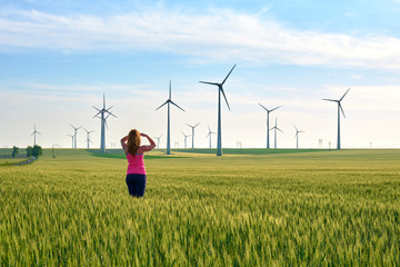 Woman and wind turbines at sunset, in a field of green rye, with warm sun light. Concept for sustainable, renewable energy.