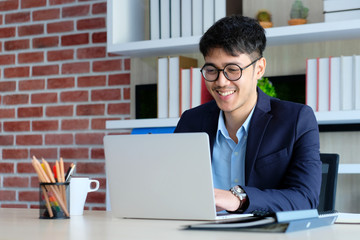 Young asian businessman smiling while working with laptop computer at office, business office lifestyle concept