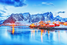 Amazing Dusk Panorama Of Picturesque Peaks Of Reine Village On Lofoten Islands Archipelago In Norway. Classical Red Rorbu - Traditional Lofoten Fishing House And And Street Lights Reflected In The Sea