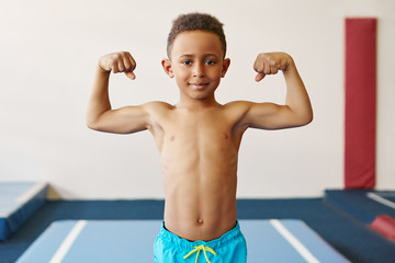 Wall Mural - Children, fitness, health and ethnicity concept. Portrait of dark skinned black boy of school age exercising at gym, preparing for competition, posing topless, demonstrating his tensed muscles