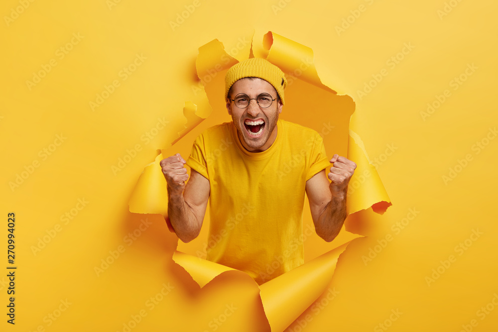 Yeah, we did it! Triumphing emotive man shouts for favourite team, yells from joy, wears yellow hat and t shirt, keeps fists clenched in victory gesture, feels upbeat, poses in torn paper hole. - obrazy, fototapety, plakaty 
