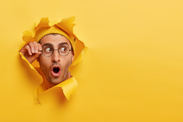 Wall Mural - Astonished unshaven man gazes through hole in yellow paper, wears round spectacles, opens mouth with surprisement, copy space to insert text or slogan. Effect of torn paper. Discount and sale