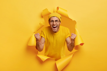 Yeah, we did it! Triumphing emotive man shouts for favourite team, yells from joy, wears yellow hat and t shirt, keeps fists clenched in victory gesture, feels upbeat, poses in torn paper hole.