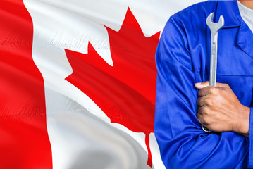 Canadian Mechanic in blue uniform is holding wrench against waving Canada flag background. Crossed arms technician.