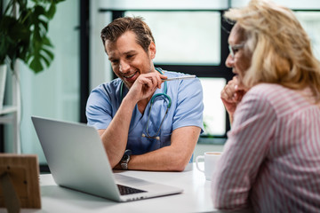 Wall Mural - Happy doctor and his patient using laptop together at medical clinic.