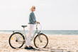 © beatleoff - Pretty blonde girl in white pants and denim coat standing on the beach with bicycle. Atrractive woman relaxing near the sea after bike ride