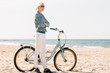 © beatleoff - Pretty blonde girl in white pants and denim coat standing on the beach with bicycle. Atrractive woman relaxing near the sea after bike ride
