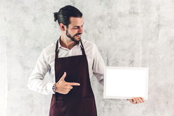 Portrait of handsome bearded barista man small business owner smiling and holding empty board wooden frame with white mockup blank in a cafe